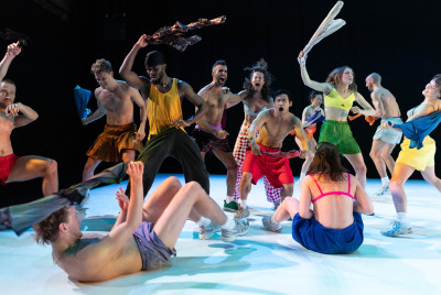 Un gruppo di dodici danzatori in abiti sportivi e colorati, mentre danzano con energia