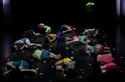 Diversi performer in abiti colorati rannicchiati a terra sullo spazio scenico