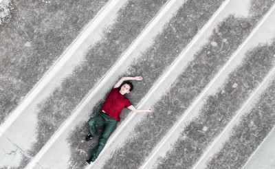 Una performer in maglia rossa e pantaloni verdi risalta distesa su un pavimento di cemento