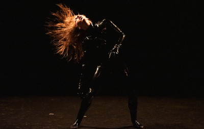 In uno spazio buio, un performer in tuta di lattice scuote la sua rossa chioma